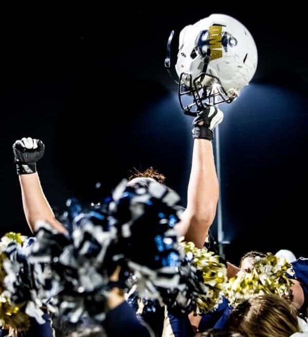 a dma football player raises his dma football helmet high in the air as he is surrounded by his happy peers